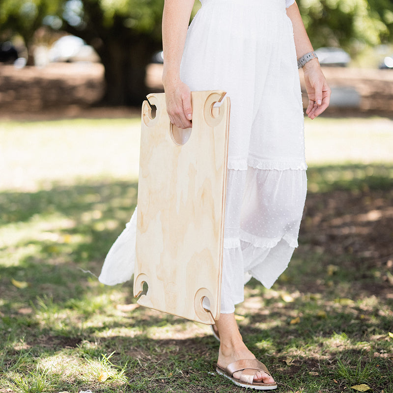 A woman in a white maxi skirt carrying a folding picnic table as she walks on green grass.