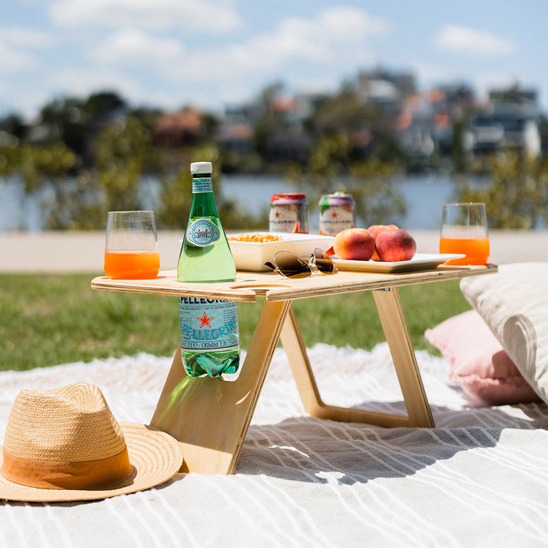 Deluxe folding picnic table sitting on a beige rug in the sunshine. On the table are peaches, drink cans and a bottle of sparkling water in the bottle anchor. A straw hat and cushions sit on the picnic rug.