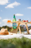 Outdoors picnic set up with blue sky. Folding picnic table and straw hat are on a picnic rug with a bottle of sparkling water, glasses and peaches on the table.