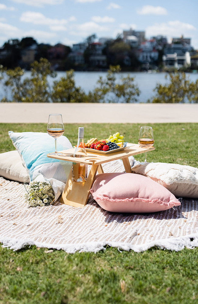 Deluxe four glass folding picnic table with a bottle of rose, fruit platter and wine glasses on a picnic rug with pink and blue cushions.