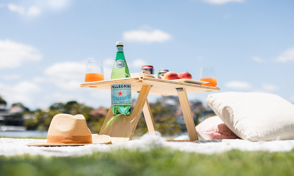 Deluxe folding picnic table sitting on a picnic rug with blue sky in the background. On top of the table are sparkling water, peaches and pink drinks. Cushions and straw hat are on the ground beside the table.