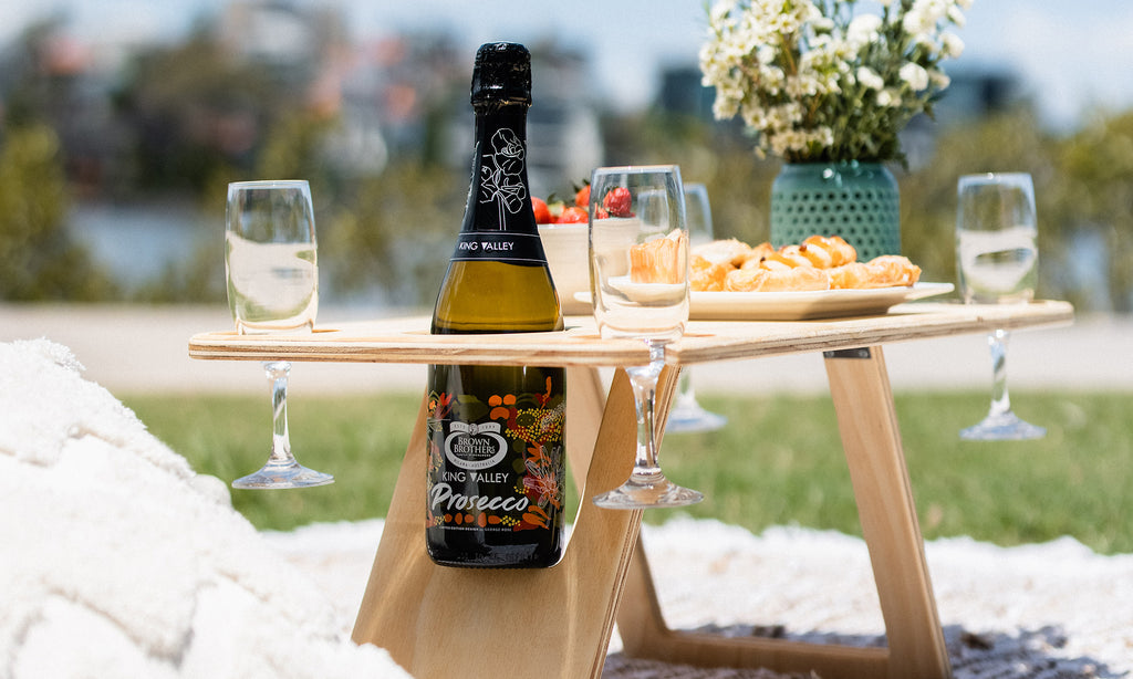 Deluxe folding picnic table with a bottle of prosecco and four champagne flutes. Pasties, strawberries and a teal vase with white flowers sit on top of the table.