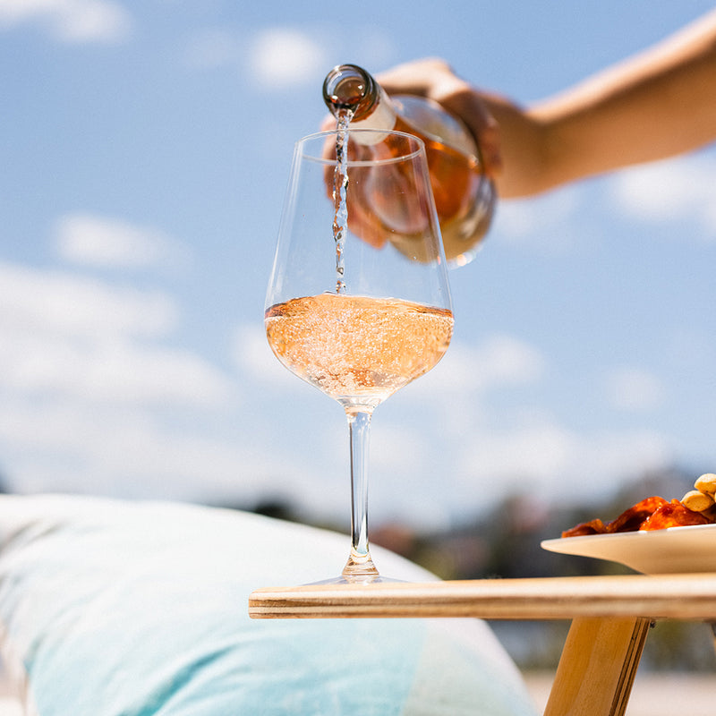 A woman's hand pouring a bottle of rose wine into a wine glass that sits on top of an Outdoors Hello folding wine table. Blue sky and blue cushion are in the background.