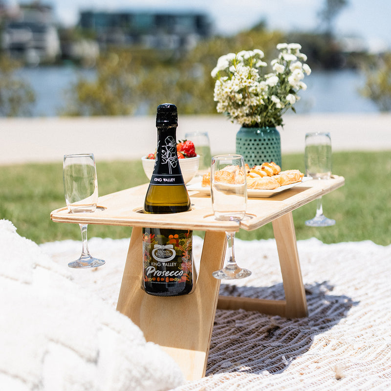 Deluxe folding picnic table sitting on a beige picnic rug holding a bottle of prosecco and four champagne flutes. On the table is a teal vase of white flowers, bowl of strawberries and plate of pastries.