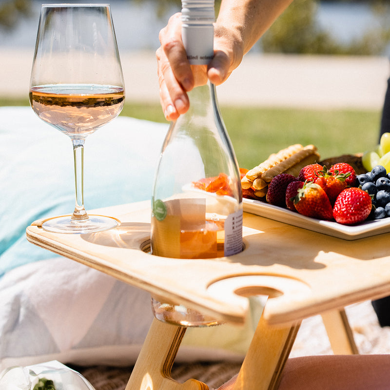 A bottle of rose being placed into the wine anchor slot on an Outdoors Hello folding picnic table. A glass of rose and fruit platter sit on top of the table. There's a blue cushion in the background.