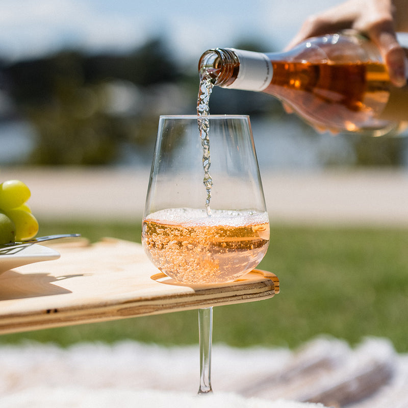 A bottle of rose wine is being poured into a wine glass that sits in a slot on the folding picnic table outdoors.
