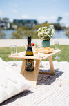 Folding picnic table on a picnic rug outdoors by the river. There's a bottle of prosecco and champagne flutes, white flowers, strawberries and pastries are on the table.
