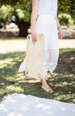 Woman in white maxi skirt carrying a folding picnic table in the park walking towards a picnic rug.
