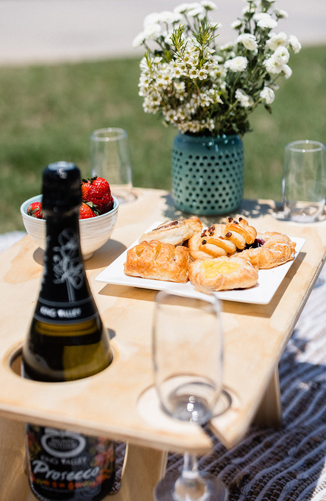 Close up image of folding wine table holding a bottle of prosecco, white flowers in a teal vase, champagne flutes, pastries and strawberries.