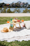 A picnic setting with a deluxe folding picnic table on a rug. A bottle of sparkling water as well as pink drink cans, pretzels and peaches are on the table. A straw hat and cushions are also sitting on the picnic rug.