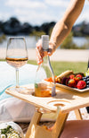 Large folding picnic table with a woman's hand placing a bottle of rose into the wine bottle holder. A glass of rose sits on the table with a fruit platter.