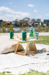 Divine folding picnic table sitting on a picnic rug with two green cans of beer, bottle of sparkling water, white bowl and two cups with green palm trees on them.