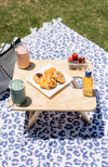A divine folding picnic table sitting on a dark blue and white leopard print rug. On top of the table are one pink and one green resuable coffee cups, a pop top apple juice bottle, plate of pastries and small container of grapes, blueberries and strawberries.