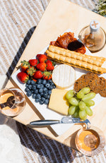 An overhead shot of a grazing platter, bottle of wine and two wine glasses on a folding picnic table sitting on a white and beige picnic rug.