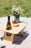 A divine folding picnic table sitting on a jute picnic rug. The table is holding a bottle of prosecco, two champagne flutes, bowl of strawberries and a teal vase with white flowers.