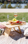 Divine two glass folding picnic table sitting on a picnic rug by the river holding a bottle of rose and wine glasses. A platter of grapes, strawberries and antipasto is sitting on the table.
