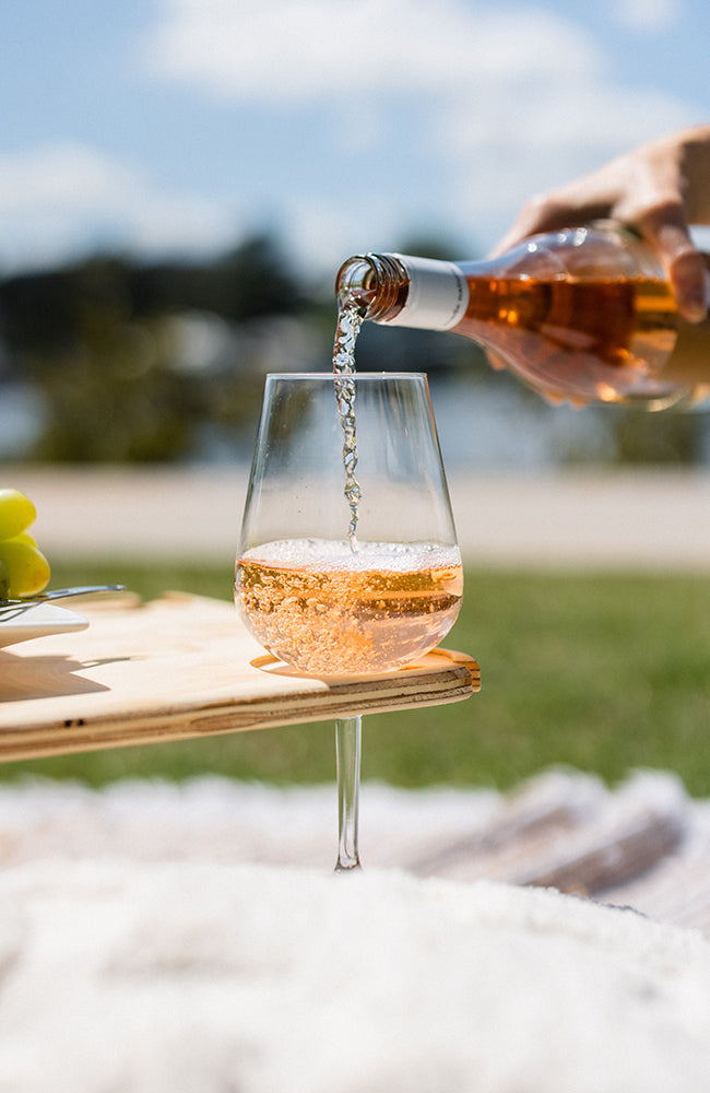 A hand pouring a bottle of rose into a wine glass on a folding picnic table.