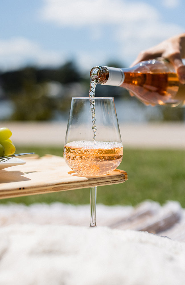 Pouring a bottle of rose into a wine glass that sits on a folding picnic table outside.