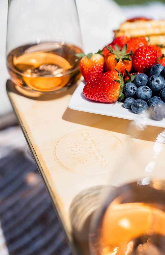 Close up of an Outdoors Hello logo on a folding picnic table. Wine glasses with rose and a plate of strawberries and blueberries are on the table.